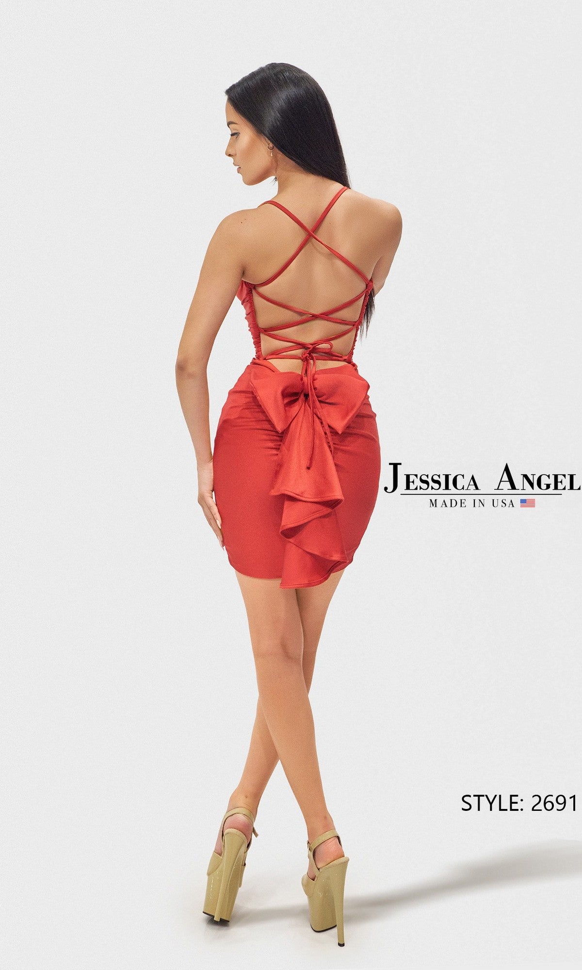 A woman stands in high heels against a plain background, wearing the Jessica Angel 2691 short prom dress—red, strappy, with crisscross open back and a large bow detail.